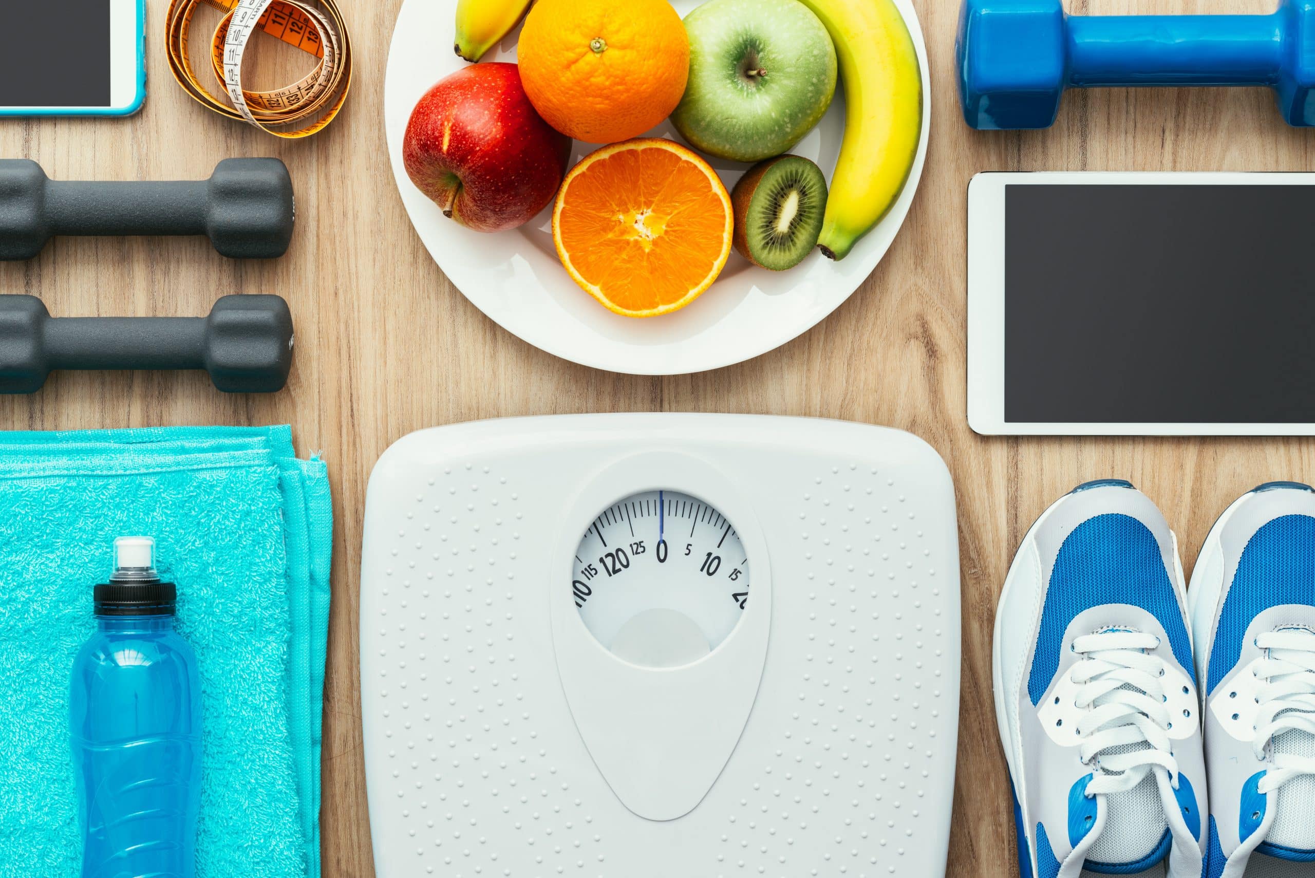 Pictured is a blue water bottle, a bright blue towel, a white scale, a pair of blue and white tennis shoes, an electronic tablet, a set of black weights, a blue weight, a plate of fruits including an orance, a red apple, a greem apple, and a banana, a cell phone, and a measuring tape. This image is included in Passive Income MDs Saturday Syndication with White Coat Investor, titled "5 Ways that Weight Loss is Like Investing."