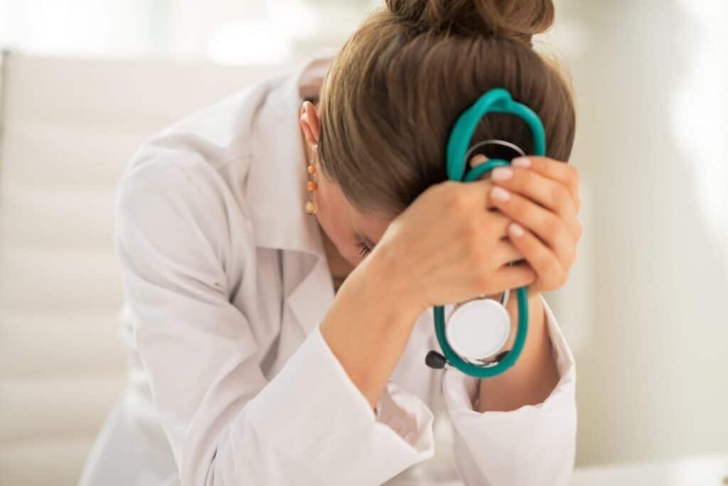 Pictured is a female physician who is head down in her hands while holding a teal green stethoscope. She apprears to be distressed and miserable. This photo is featured on the Passive Income MD blog, where we have featured The Physician Philosopher in a blog post titled, "I Am a Doctor and I Hate My Job."