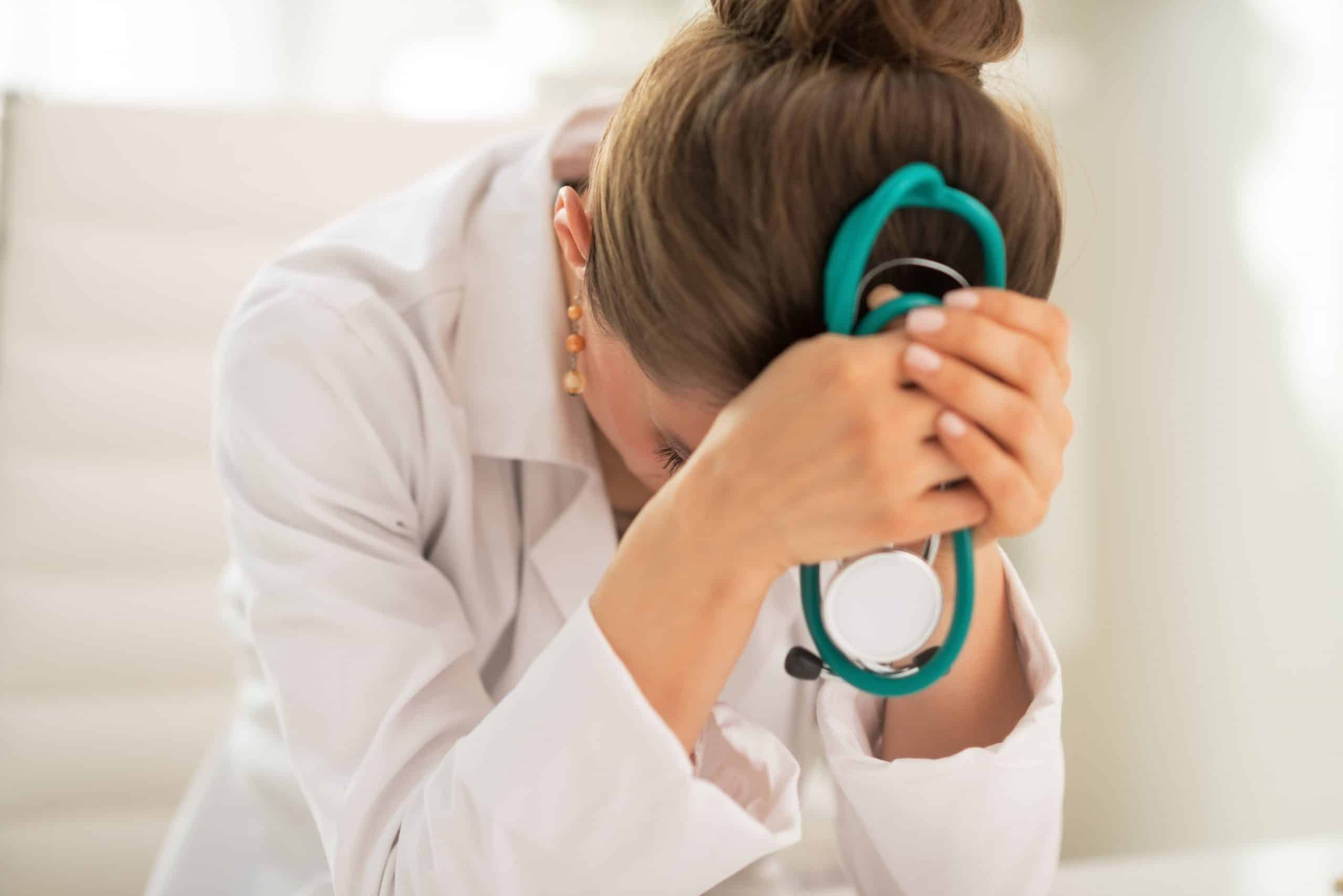 Pictured is a female physician who is head down in her hands while holding a teal green stethoscope. She apprears to be distressed and miserable. This photo is featured on the Passive Income MD blog, where we have featured The Physician Philosopher in a blog post titled, "I Am a Doctor and I Hate My Job."